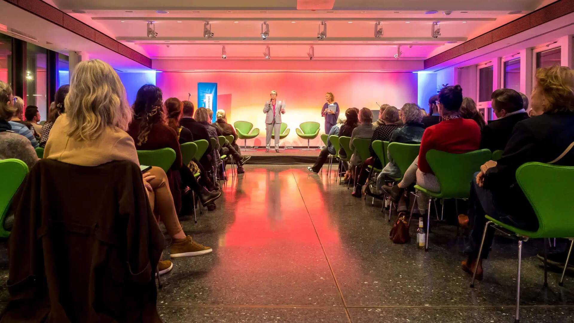 Unser Jubiläum 2023 Veranstaltungssaal, in dem Menschen vor einer Bühne sitzen. Auf der Bühne zwei Rednerinnen.