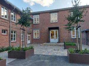 Eingang zu einem Klinker-Gebäude mit weißer Tür, vor dem Eingang ist ein Platz mit Baumpflanzung. Das Gebäude geht links in eine weiteren Gebäudeteil über.