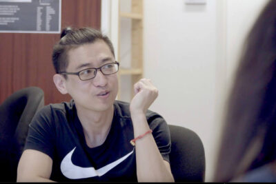 Ein junger Mann asiatischer Herkunft mit Brille, Kurzarm-Shirt und zurückgebundenen Haaren sitzt an einem Tisch und schaut zur Seite im Gespräch mit einer oder mehreren Personen